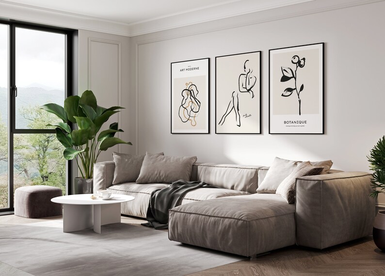 May include: A living room interior with a large sectional sofa in a light beige color. The sofa is positioned in front of a white wall with three framed black and white line art prints. The prints feature abstract shapes, a figure, and a botanical illustration. The text on the prints reads "Art Moderne" and "Botanique". A large potted plant with green leaves is in the corner of the room. A white coffee table with a unique base design sits in front of the sofa. A small gray ottoman is in the foreground.