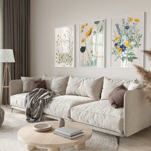 May include: A living room scene featuring a beige sofa with throw pillows and a blanket. Three framed floral art prints hang above the sofa. A wooden floor lamp, round coffee table, and decorative vases complete the room's decor.
