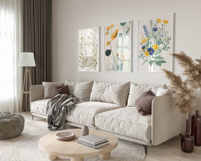 May include: A living room scene featuring a beige sofa with throw pillows and a blanket. Three framed floral art prints hang above the sofa. A wooden floor lamp, round coffee table, and decorative vases complete the room's decor.