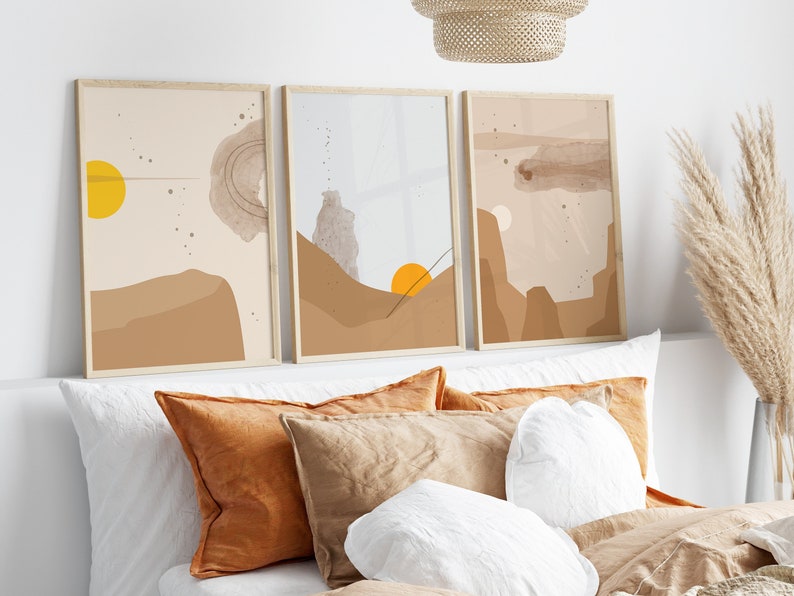 May include: Three framed abstract art prints with a desert landscape theme. The prints feature a muted color palette of beige, brown, and yellow. The prints are hung above a bed with white bedding and brown and orange pillows.