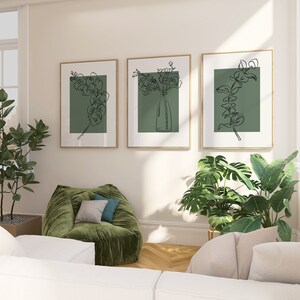May include: Three framed line drawings of flowers and greenery on a white wall. The drawings are in black and white and have a green background. The frames are a light wood colour. There is a green velvet armchair in the foreground.