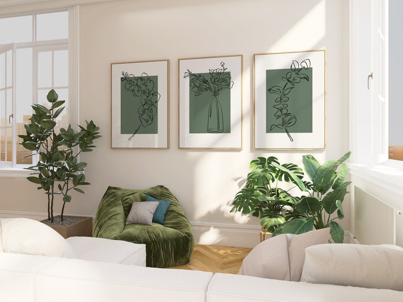 May include: Three framed line drawings of flowers and greenery on a white wall. The drawings are in black and white and have a green background. The frames are a light wood colour. There is a green velvet armchair in the foreground.