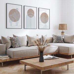 May include: A light beige sectional sofa with a wooden frame and a matching coffee table. The sofa has several pillows, including a patterned pillow. Three framed abstract prints with brown and blue geometric designs hang on the wall behind the sofa.