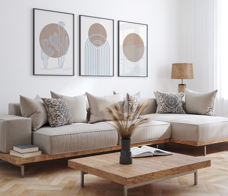May include: A light beige sectional sofa with a wooden frame and a matching coffee table. The sofa has several pillows, including a patterned pillow. Three framed abstract prints with brown and blue geometric designs hang on the wall behind the sofa.