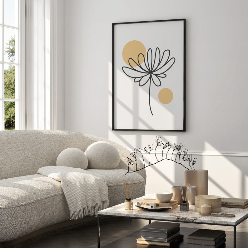 May include: A living room interior with a white couch, a coffee table, and a black framed print with a line drawing of a flower and two yellow circles. The coffee table has a marble top and is decorated with a vase, candles, and books.