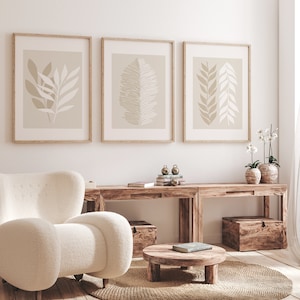 May include: Three framed prints with beige backgrounds and abstract leaf designs hang on a white wall above a wooden console table. The table has two sections and is decorated with books, decorative objects, and two potted plants. A white armchair sits in front of the table.