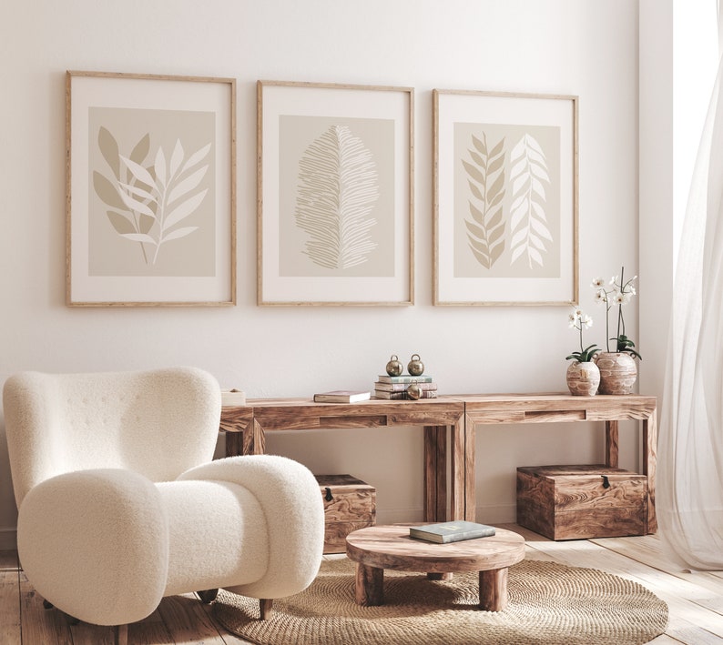 May include: Three framed prints with beige backgrounds and abstract leaf designs hang on a white wall above a wooden console table. The table has two sections and is decorated with books, decorative objects, and two potted plants. A white armchair sits in front of the table.