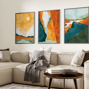 May include: Three abstract landscape paintings in a living room setting. The paintings are framed in gray and feature a mix of orange, yellow, blue, and green colors. The paintings are hung on a white wall above a beige sectional sofa with pillows and a throw blanket.