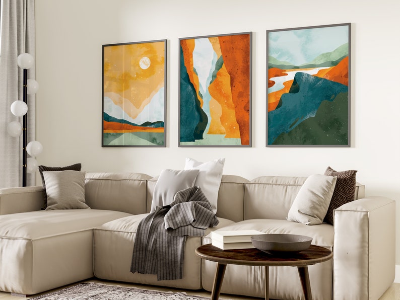 May include: Three abstract landscape paintings in a living room setting. The paintings are framed in gray and feature a mix of orange, yellow, blue, and green colors. The paintings are hung on a white wall above a beige sectional sofa with pillows and a throw blanket.