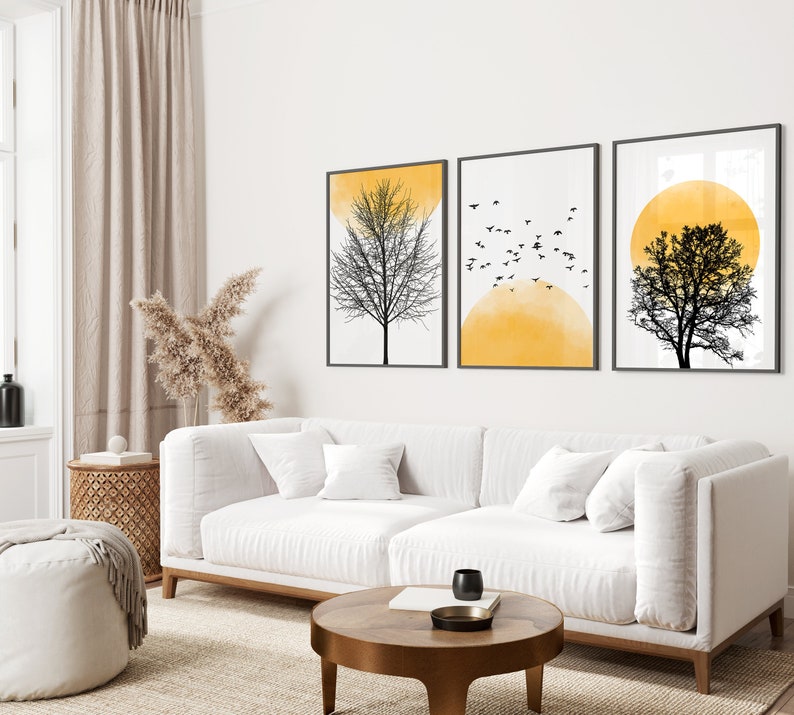 May include: Three black and white minimalist art prints featuring silhouettes of trees and a yellow sun. The prints are framed and hung on a white wall above a white couch with pillows.