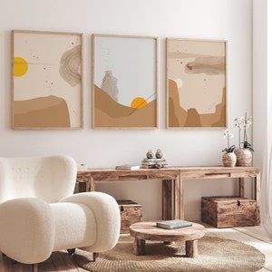 May include: Three abstract landscape prints in brown, beige, and blue tones with a yellow sun in the top left corner of the leftmost print. The prints are framed in natural wood and hung on a white wall above a wooden console table with two wooden storage boxes.