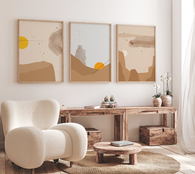 May include: Three abstract landscape prints in brown, beige, and blue tones with a yellow sun in the top left corner of the leftmost print. The prints are framed in natural wood and hung on a white wall above a wooden console table with two wooden storage boxes.