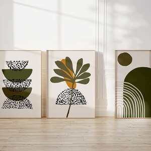 May include: Three framed abstract art prints with a minimalist design featuring green, brown, and white colors. The prints feature geometric shapes, a stylized plant, and a circle.