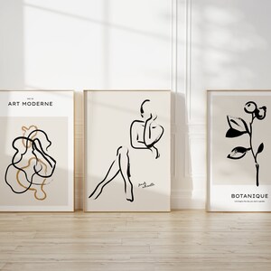 May include: Three framed art prints with black and white line drawings. The left print has a black and gold abstract design with the text "ART MODERNE" at the top. The middle print has a black line drawing of a person sitting with the text "Femme Silhouette" at the bottom. The right print has a black line drawing of a plant with the text "BOTANIQUE" at the bottom.