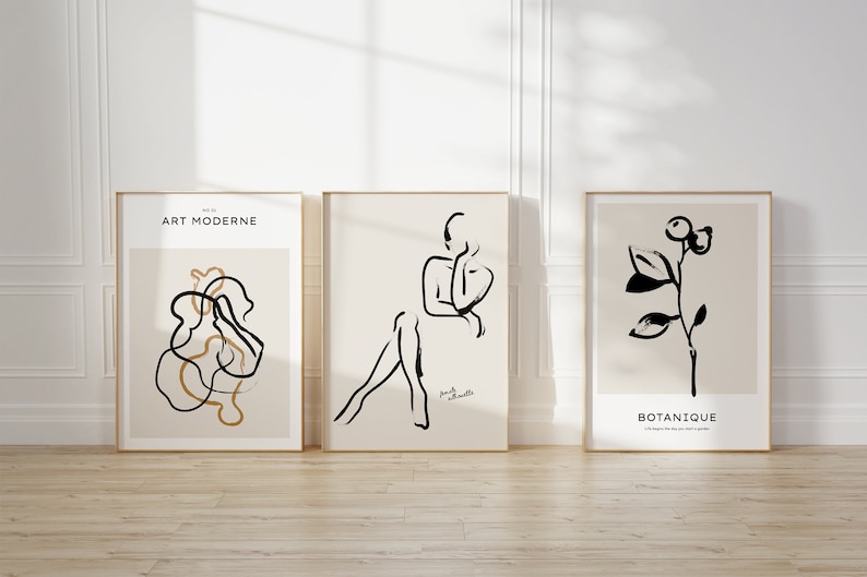 May include: Three framed art prints with black and white line drawings. The left print has a black and gold abstract design with the text "ART MODERNE" at the top. The middle print has a black line drawing of a person sitting with the text "Femme Silhouette" at the bottom. The right print has a black line drawing of a plant with the text "BOTANIQUE" at the bottom.
