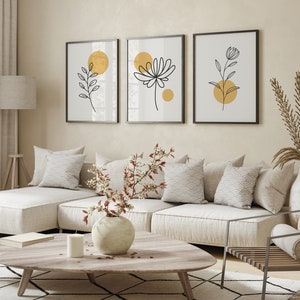 May include: Three minimalist black and white line art prints with gold circles on a white background. The prints feature stylized floral and leaf designs. The prints are hanging on a beige wall above a white sectional sofa with pillows.