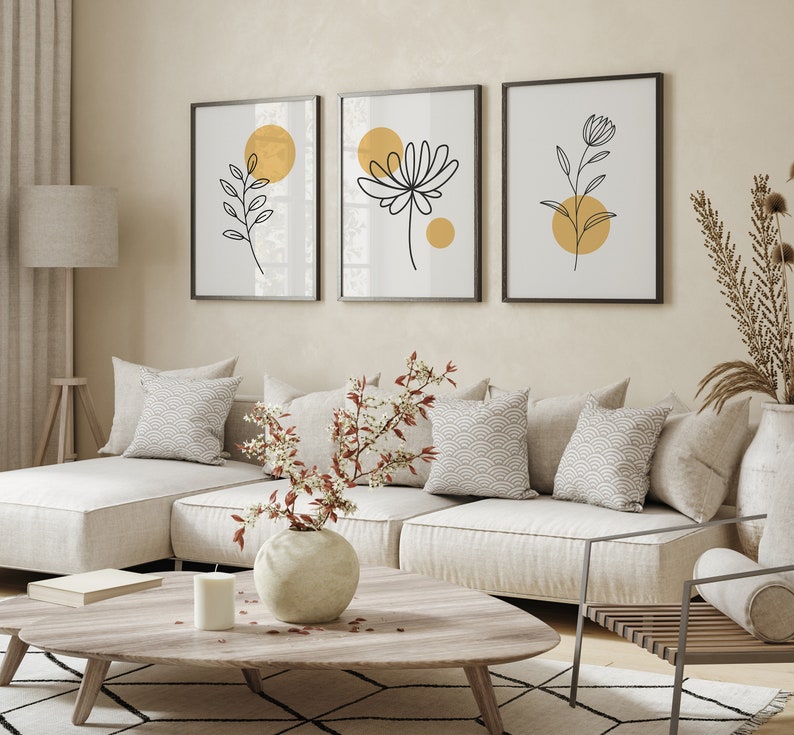 May include: Three minimalist black and white line art prints with gold circles on a white background. The prints feature stylized floral and leaf designs. The prints are hanging on a beige wall above a white sectional sofa with pillows.