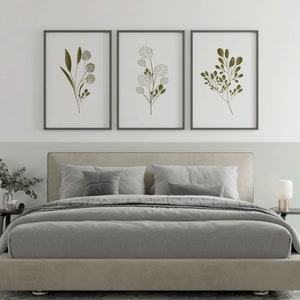 May include: Three framed botanical prints featuring minimalist line drawings of green leaves and white flowers on a white background. The prints are hanging on a white wall above a grey upholstered bed with white pillows.
