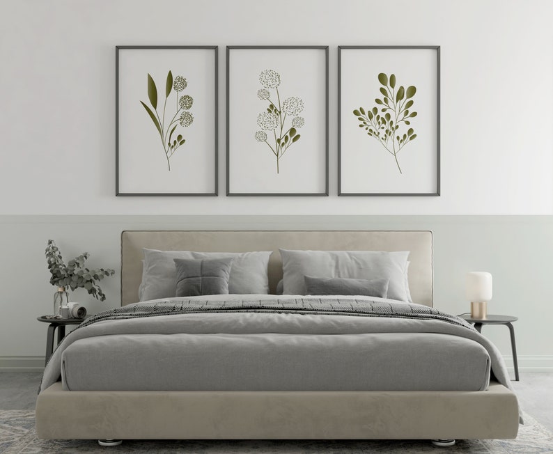 May include: Three framed botanical prints featuring minimalist line drawings of green leaves and white flowers on a white background. The prints are hanging on a white wall above a grey upholstered bed with white pillows.
