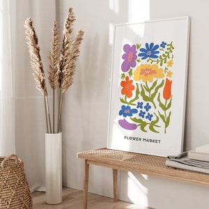K&ouml;nnte beinhalten: Ein wei&szlig;er gerahmter Druck mit einer bunten Illustration von Blumen und dem Text "FLOWER MARKET". Der Druck h&auml;ngt an einer wei&szlig;en Wand &uuml;ber einer Holzbank.