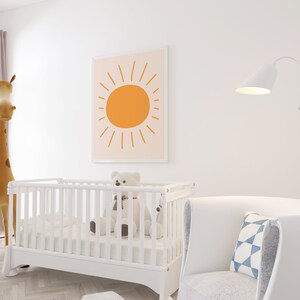 May include: A nursery scene featuring a white crib with a teddy bear, a framed print of an orange sun, a giraffe plush toy, and a white armchair with a blue and white patterned pillow. A white floor lamp is also visible.