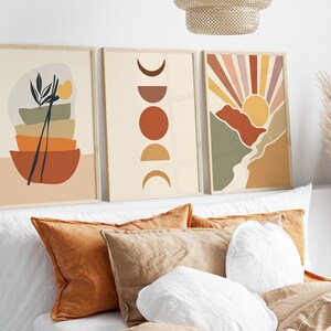 May include: Three framed abstract art prints with a minimalist design. The prints feature geometric shapes in shades of brown, orange, yellow, and green. The prints are hung on a white wall above a bed with pillows.