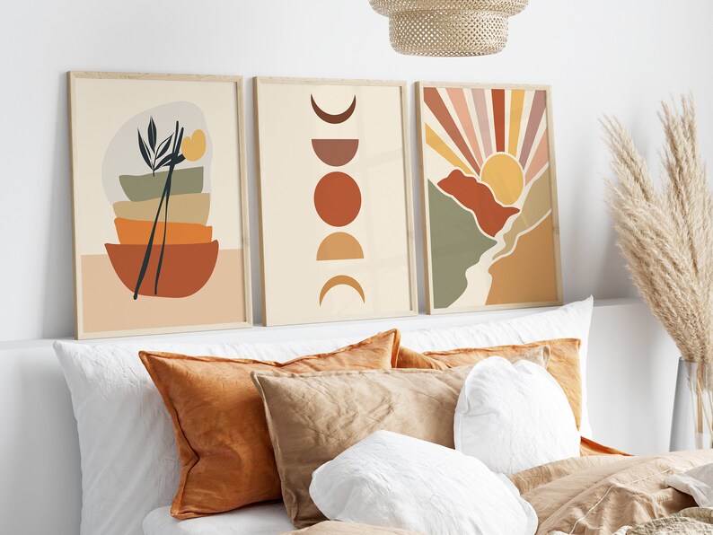May include: Three framed abstract art prints with a minimalist design. The prints feature geometric shapes in shades of brown, orange, yellow, and green. The prints are hung on a white wall above a bed with pillows.
