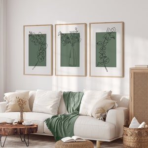 May include: Three framed prints featuring line drawings of flowers and foliage on a sage green background. The prints are displayed on a white wall above a white sofa with a green throw blanket.