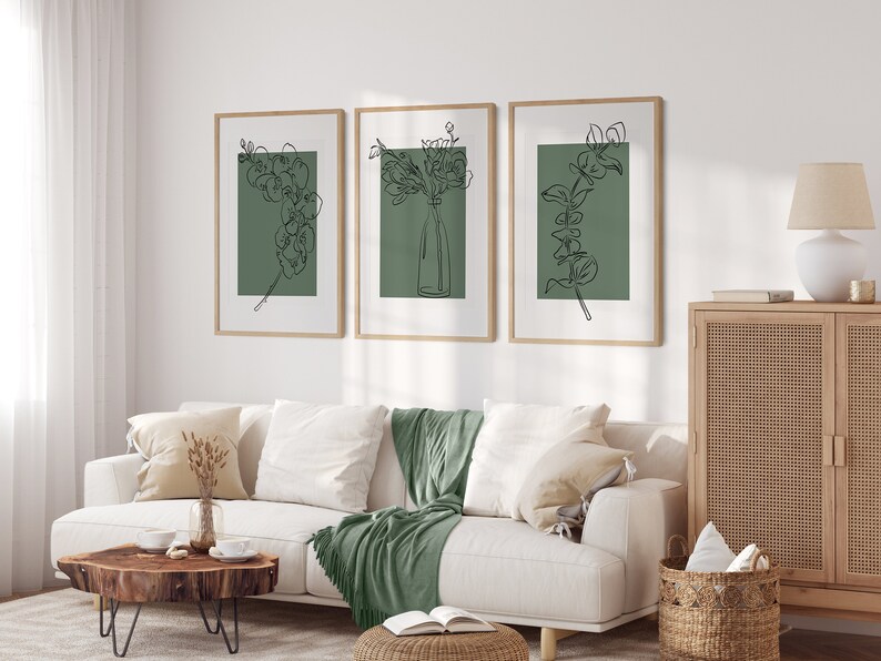 May include: Three framed prints featuring line drawings of flowers and foliage on a sage green background. The prints are displayed on a white wall above a white sofa with a green throw blanket.