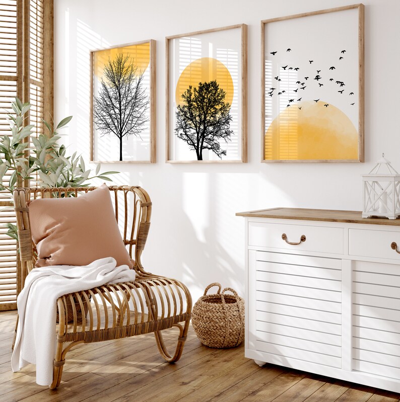 May include: A white cabinet with two drawers and two doors, a wicker chair with a brown cushion, a woven basket, and three framed prints with black silhouettes of trees and yellow circles.