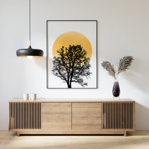 May include: A black and white print of a tree with a yellow circle behind it hangs above a light wood cabinet with slatted doors. A vase with dried flowers sits on top of the cabinet.