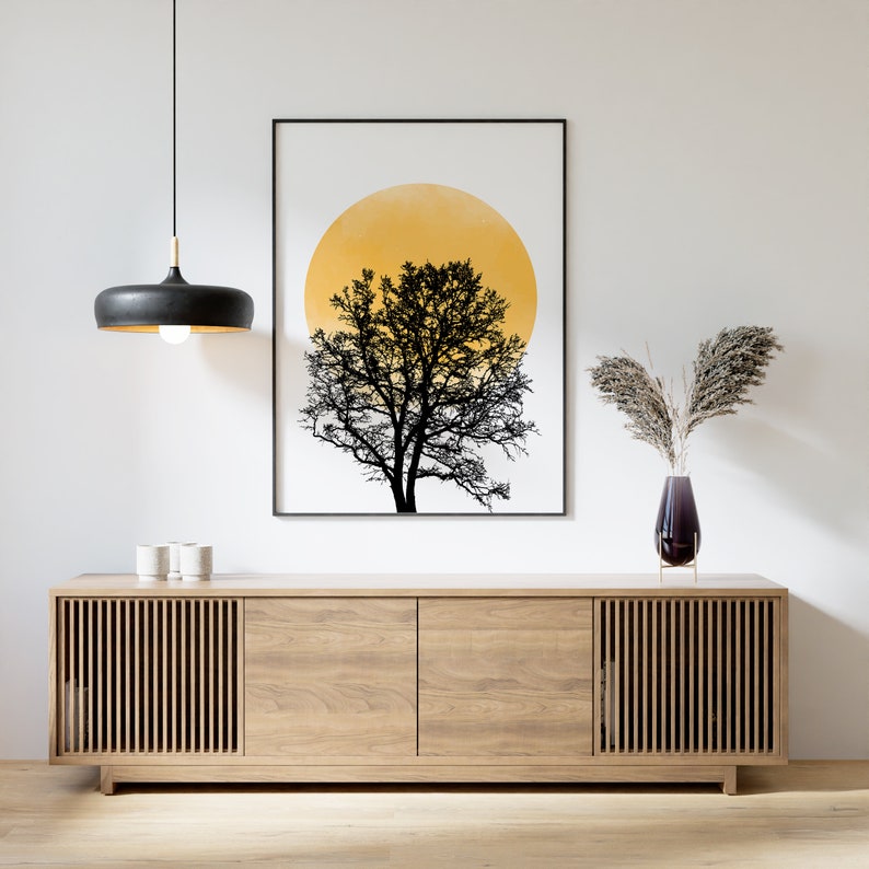 May include: A black and white print of a tree with a yellow circle behind it hangs above a light wood cabinet with slatted doors. A vase with dried flowers sits on top of the cabinet.
