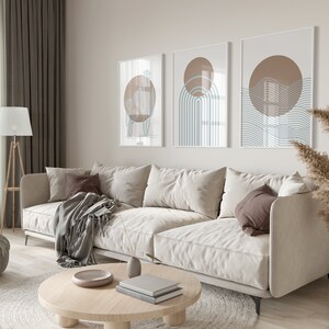 May include: A light beige sectional sofa with a brown throw blanket and pillows in a living room with a coffee table, a lamp, and a vase of dried pampas grass. Three abstract art prints with brown and blue geometric shapes hang on the wall.