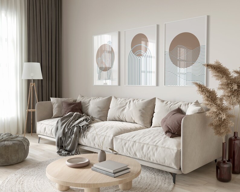 May include: A light beige sectional sofa with a brown throw blanket and pillows in a living room with a coffee table, a lamp, and a vase of dried pampas grass. Three abstract art prints with brown and blue geometric shapes hang on the wall.