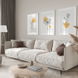 May include: A white sectional sofa with brown pillows sits in a living room with a wooden coffee table and a rug. Three framed prints with black line drawings of flowers and leaves with yellow circles hang on the wall.