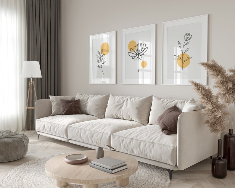 May include: A white sectional sofa with brown pillows sits in a living room with a wooden coffee table and a rug. Three framed prints with black line drawings of flowers and leaves with yellow circles hang on the wall.