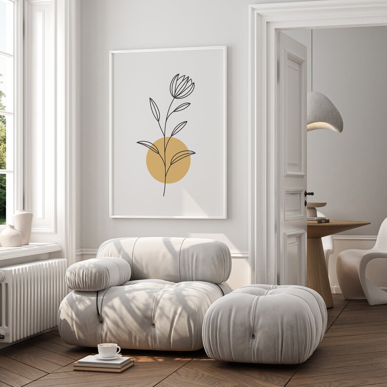 May include: A white, tufted armchair with a matching ottoman. The armchair has a single armrest and a large, square seat cushion. The ottoman is round and has a similar tufted design. A framed print of a black line drawing of a flower with a yellow circle behind it hangs on the wall behind the armchair.
