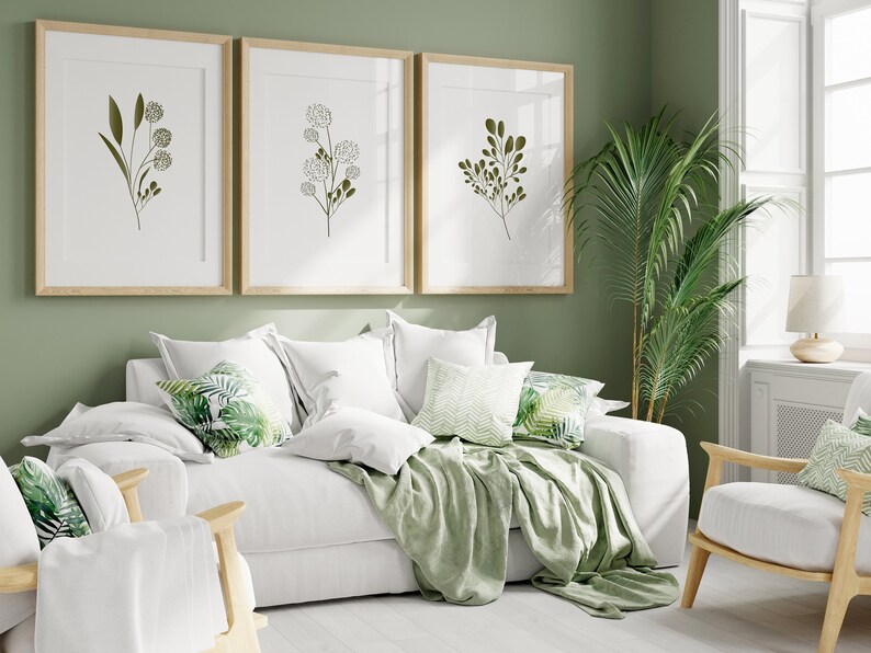 May include: A white sofa with green throw cushions and a green throw blanket. There are three framed prints of green botanical illustrations on the wall behind the sofa. A green potted plant is in the corner of the room.