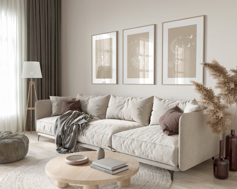 May include: A light beige sectional sofa with a gray throw blanket and brown pillows sits in a living room with a round wooden coffee table and three framed prints of dandelion flowers on the wall.