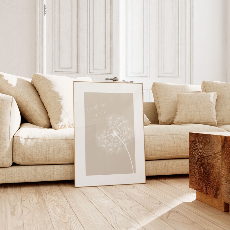 May include: A framed print of a dandelion with its seeds blowing in the wind. The print is on a beige background and has a white frame. The print is sitting on a wooden floor in front of a beige couch.