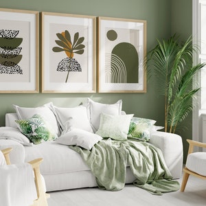 May include: A white sofa with green throw pillows and a green throw blanket. The sofa is in front of a green wall with three framed abstract art prints. A green potted plant is in the corner of the room.