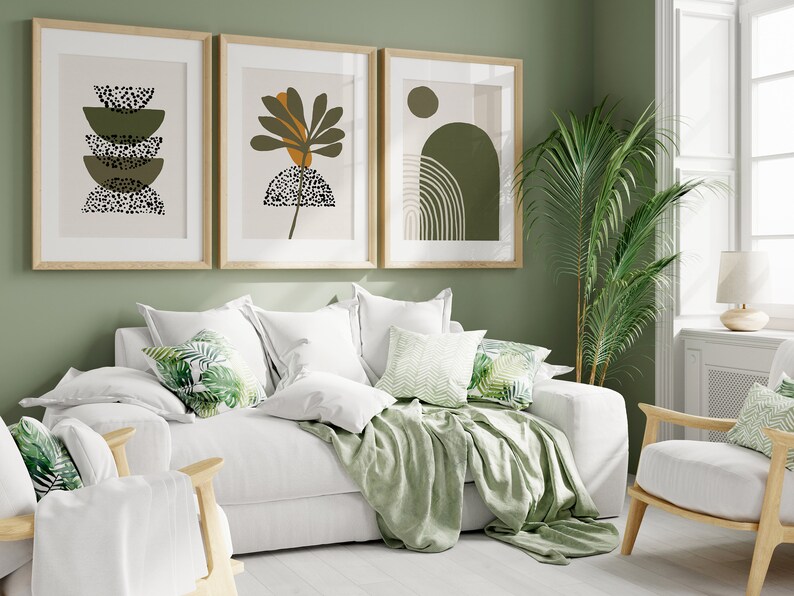 May include: A white sofa with green throw pillows and a green throw blanket. The sofa is in front of a green wall with three framed abstract art prints. A green potted plant is in the corner of the room.