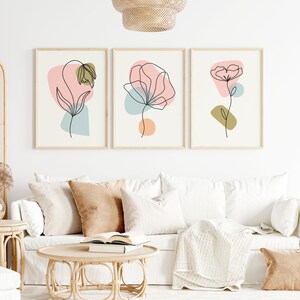 May include: Three minimalist line art prints of flowers with abstract shapes in pink, blue, and green colors. The prints are framed in natural wood and hung on a white wall above a white couch.
