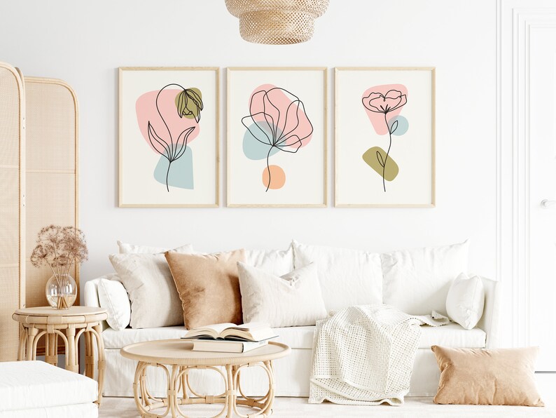 May include: Three minimalist line art prints of flowers with abstract shapes in pink, blue, and green colors. The prints are framed in natural wood and hung on a white wall above a white couch.