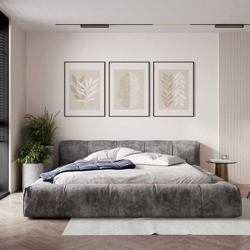 May include: A gray upholstered bed with a dark blue blanket and three framed prints of stylized leaves on a white wall.