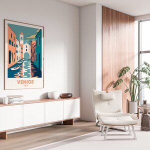 May include: A white armchair with a footrest sits in front of a large window with a view of a city. A wooden cabinet with white doors is to the left of the chair. A framed poster with a colorful illustration of Venice, Italy, hangs on the wall above the cabinet. The poster reads "Venice, Italy".