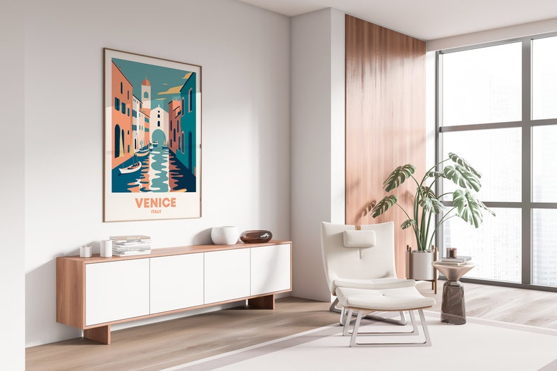 May include: A white armchair with a footrest sits in front of a large window with a view of a city. A wooden cabinet with white doors is to the left of the chair. A framed poster with a colorful illustration of Venice, Italy, hangs on the wall above the cabinet. The poster reads "Venice, Italy".