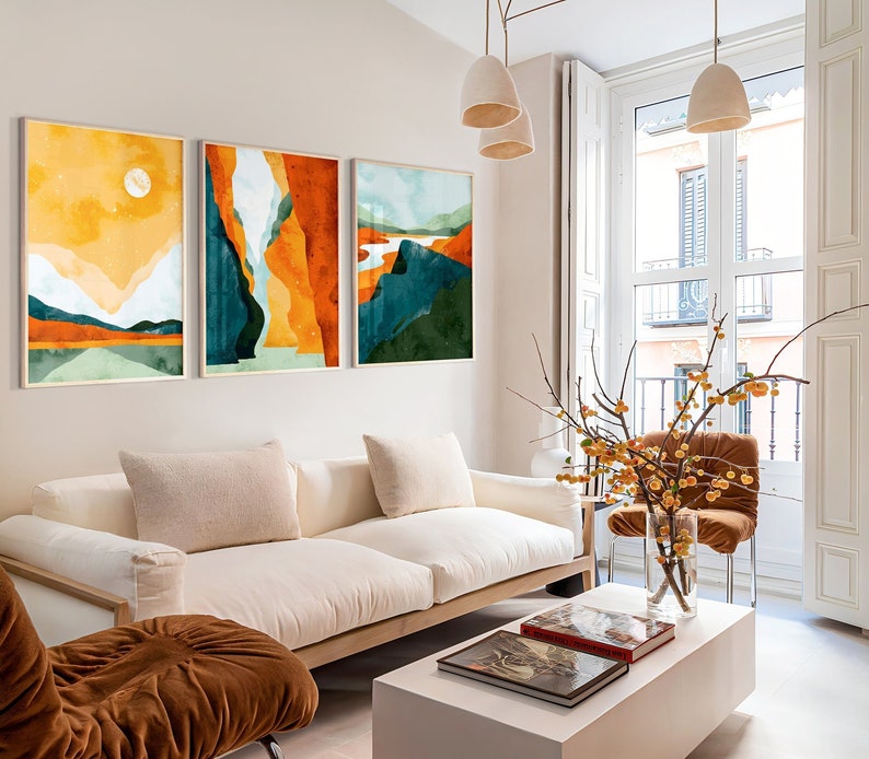 May include: Three abstract landscape paintings in a living room with a white couch, a brown armchair, and a white coffee table. The paintings are in shades of yellow, orange, green, and blue.
