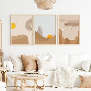May include: Three abstract landscape prints in brown, beige, and gray tones with yellow sun accents. The prints are framed in natural wood and hung on a white wall above a white couch.