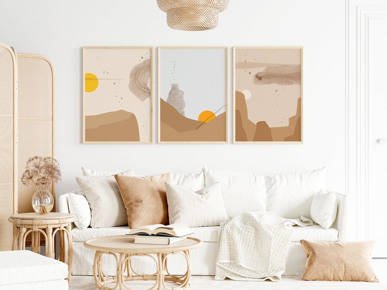 May include: Three abstract landscape prints in brown, beige, and gray tones with yellow sun accents. The prints are framed in natural wood and hung on a white wall above a white couch.